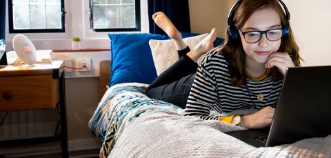 A young person in a bedroom watching laptop with headphones