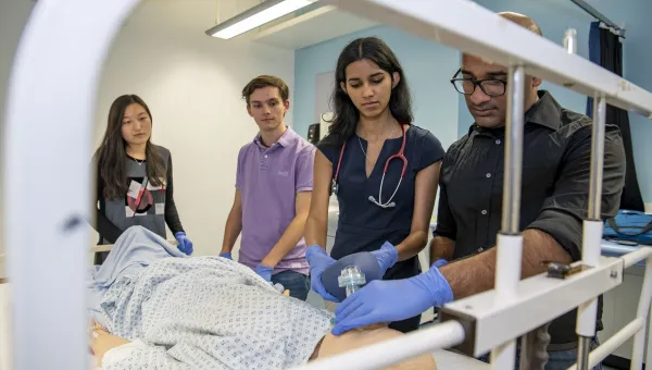A group of students watch as one of them works on a dummy patient in a hospital bed
