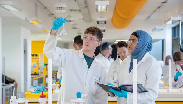 Two students wearing lab coats and protective glasses conduct an experiment. One holds up a glass beaker, while the other takes notes. They are in a laboratory, surrounded by a variety of chemistry equipment.