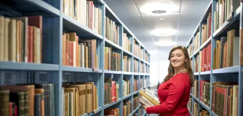 A student smiling over her shoulder as she carries a pile of books through the stacks in Hartley library.