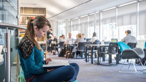 A student sits cross-legged reading a book in a bright, airy library. Behind her are shelves of books and other students working at desks.