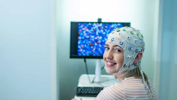 A student sits in front of a computer and turns to smile towards the camera. She is wearing a head cap that is connected to electrodes to monitor brain activity.