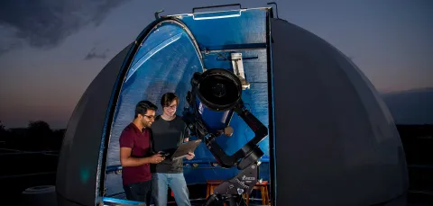A small, domed rooftop observatory opens onto a starry night sky. Inside the observatory, two students use a telescope and a laptop to collect data.