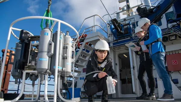 Three students outside on the deck of a boat, on a sunny day. One crouches down to collect a water sample from a large piece of technical equipment.
