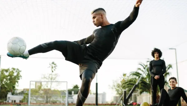 A young male footballer raising his leg parallel to the ground, to kick a ball towards the camera.