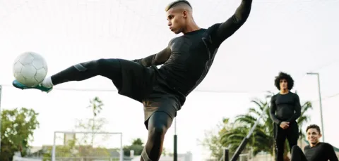 A young male footballer raising his leg parallel to the ground, to kick a ball towards the camera.