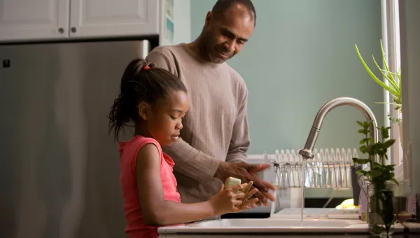 A father smiling down at his young daughter as she washes her hands carefully with soap.