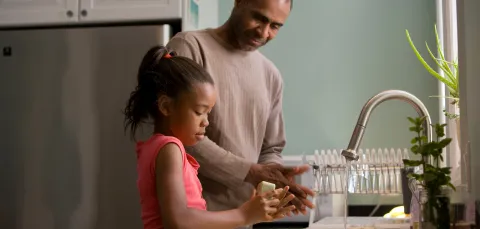 A father smiling down at his young daughter as she washes her hands carefully with soap.