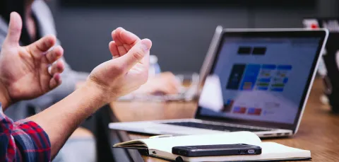 Laptop on a table with notepad and phone, user's hands in foreground 