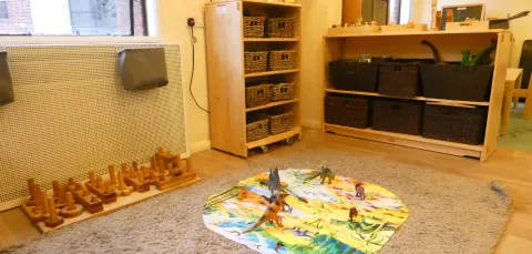 The toddler room at our Early Years Centre. A mat is laid out on the carpet with several model dinosaurs, and more toys in baskets on shelves in the background.