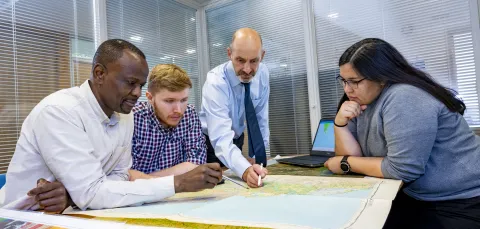 A Southampton Geospatial researcher talks with team members, standing over a map, with a laptop with a data visualisation to the side
