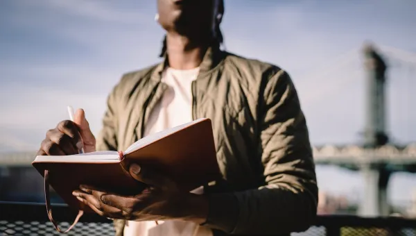 A Black man writes on a notepad as he stands outside, a blue sky and a bridge behind 