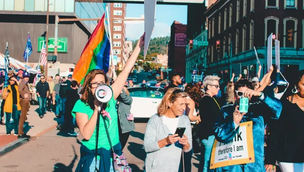 A diverse group of protestors, mainly women closest to us, hold up banners and a pride flag. One in the foreground is shouting into a loudspeaker.