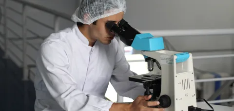 A researcher in a white lab coat looks through a microscope.