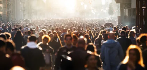 A large crowd of people spans from the foreground to the background. The people closer to us are picked out as indviduals because of a low, bright sunlight.