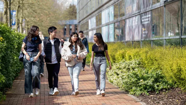A group of students of different ethnicities walk through a green, sunlit university campus, turned towards each other, in discussion