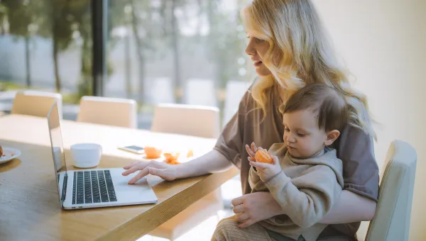 A woman sitting at a table using a laptop with one hand and a child sitting on her knee eating an orange 