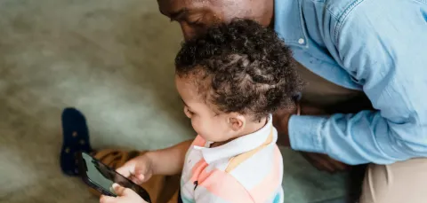 Young child playing on a phone as an adult watches them over their shoulder looks 