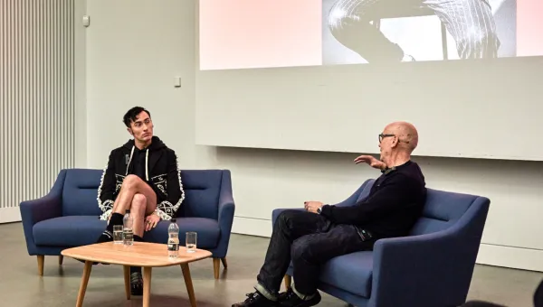 Charles Jeffrey and Professor Jonathan Faiers sit on blue sofas with a small table between them. Jonathan is facing away from the camera and has an arm raised as he speaks. Charles is facing Jonathan and appears to be concentrating on what Jonathan is saying. There is a projector behind them showing a person wearing a pinstripe suit.