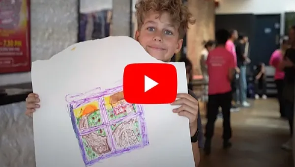 A boy holding a white banner with a drawing showing a view out of a window with the sun and green grass 
