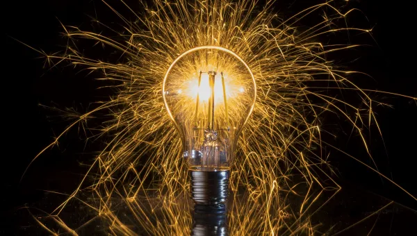 An illuminated light bulb with a vibrant shower of golden sparks radiating around it, resting on a reflective surface with a visible reflection below.