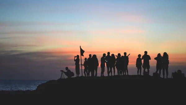A group of people as silhouettes against a colorful sunset sky, some taking photos and holding a flag, on a rocky lookout.