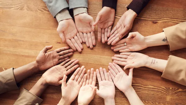 Several hands with varying skin tones turned palm-up in a circle on a wooden table, symbolizing diversity and unity.