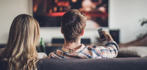 A couple sitting on a sofa, watching a wall mounted TV. The man is pointing the remote at the screen.
