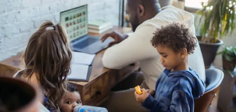 Family at home with father working on laptop, mother feeding baby, and young child playing nearby.