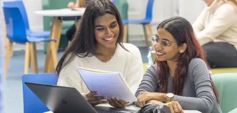 Two students huddle around a laptop together, whilst reading information on a large notepad.