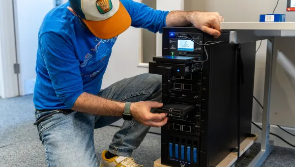 A person is inserting or removing a hard drive from a multi-bay server or data storage system in a tech workspace. They are wearing a blue shirt, jeans, and a cap with the label "Outdoor."