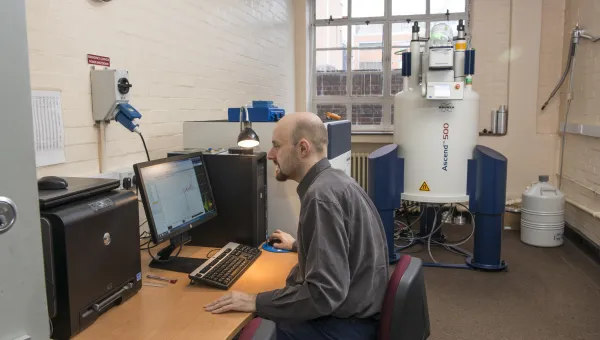 A man working  on a computer in a room with a Nuclear Magnetic Resonance machine in the background