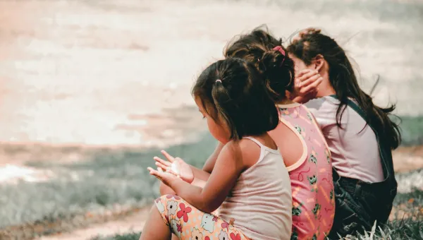 Three children sitting on grass, facing away, with colorful clothing.