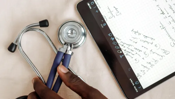 Hands write notes on a tablet with a stethoscope and a laptop near by.