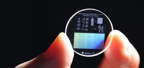 Zoomed in view of a hand holding a 5D memory crystal between two fingers. The memory crystal is a small clear disc with diagrams of human DNA on the surface.