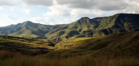 A landscape of rolling hills with long grass in the Lesotho Highlands. There is a mountain range on the horizon, and the sky is mainly cloudy with some clear blue areas.
