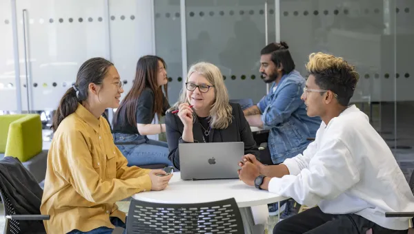 Two students and an advisor talking around a table. The advisor has a laptop open on the desk. There are other people sitting around tables in the background.