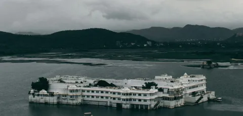 The Taj Lake Palace in Udaipur, Rajasthan, India. It is a rainy day and there are dark clouds surrounding the hills in the distance.