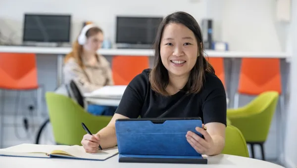 Jessie is sitting at a desk smiling at the camera. She has a tablet in 1 hand and a pen and note book in the other hand.