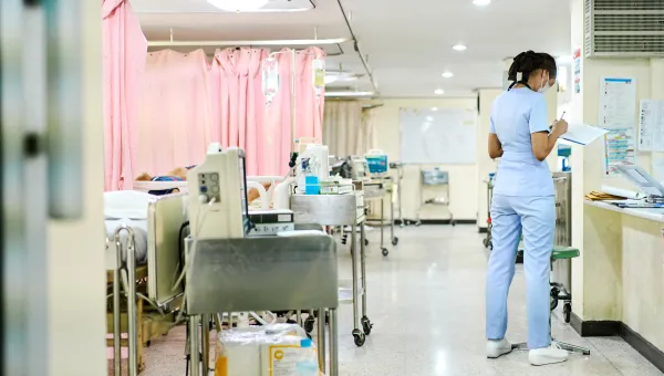 A hospital ward showing a trolley of equipment at the end of each bed. There is 1 member of staff facing away from the camera completing a chart.