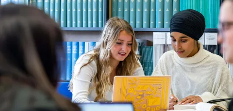 Two students discussing work in a library with a yellow laptop.