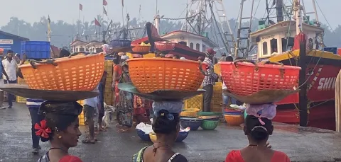 Three women at a ship harbour carrying baskets of fish on their heads.