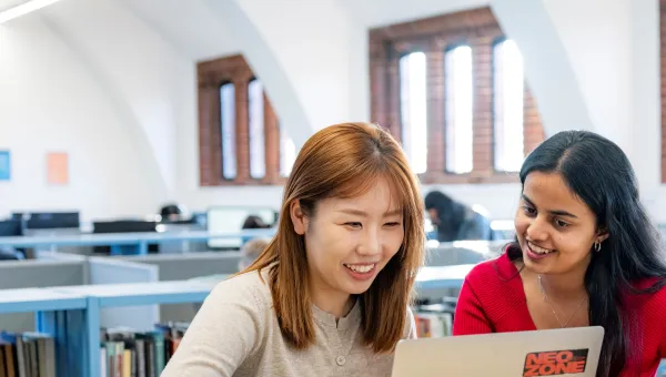 Two students sitting at a table in a library. They are smiling and looking at a laptop.