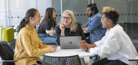 A group of three people sat around a table. They're talking in the group with a share laptop in the middle.