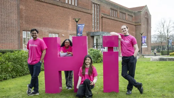 Four students leaning on large pink letters spelling Hi on the grass outside the Hartley library.
