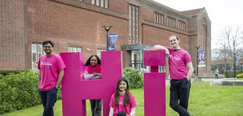 Four students leaning on large pink letters spelling Hi on the grass outside the Hartley library.