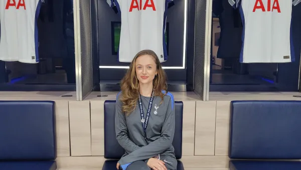 Emma Green wearing a Tottenham Hotspur tracksuit, is sitting on a chair with 3 Tottenham Hotspur shirts hanging behind her.