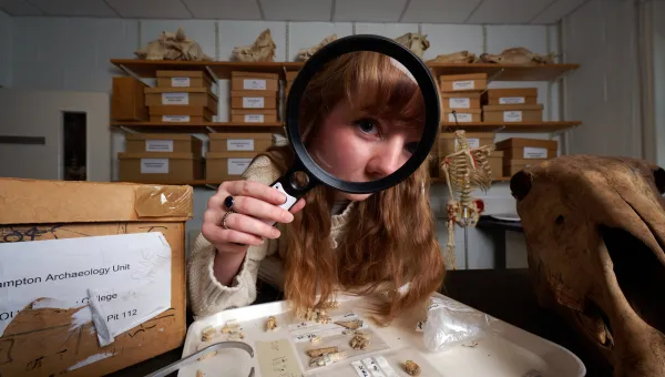 An archaeology student in an archives room, using a magnifying glass to inspect a tray of bones and fossils. The student has red hair and is wearing a cream-coloured knitted sweater. There are more fossils and bones in the background, as well as shelves filled with boxes.