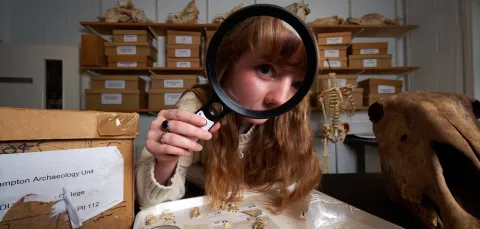 An archaeology student in an archives room, using a magnifying glass to inspect a tray of bones and fossils. The student has red hair and is wearing a cream-coloured knitted sweater. There are more fossils and bones in the background, as well as shelves filled with boxes.