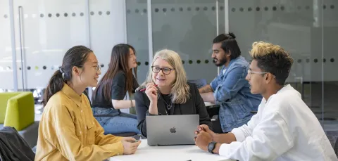 Three people sat a desk talking. The person in middle has a laptop open on the desk.